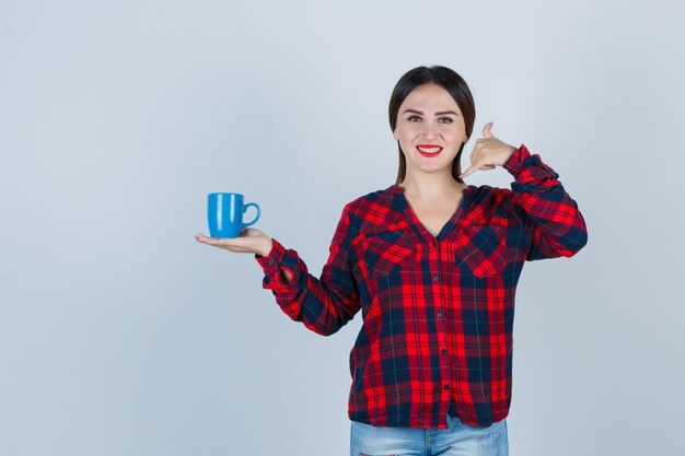 Jeune belle femme en chemise décontractée, jeans tenant une tasse tout en faisant un geste téléphonique et en ayant l'air joyeux, vue de face.