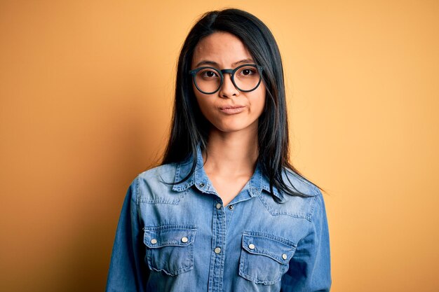 Jeune belle femme chinoise portant une chemise en jean décontractée sur fond jaune isolé, l'air endormi et fatigué, épuisé par la fatigue et la gueule de bois, les yeux paresseux le matin