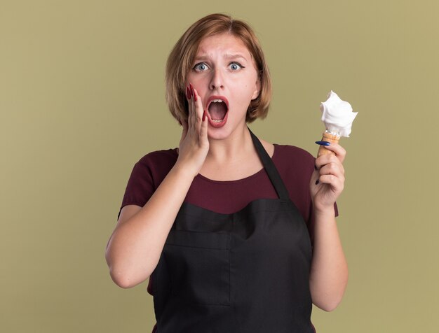 Jeune belle femme coiffeur en tablier tenant un blaireau avec de la mousse à raser à l'avant étonné et inquiet debout sur le mur vert