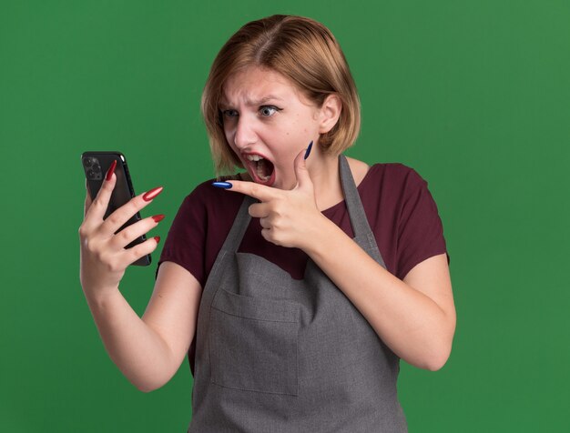 Jeune belle femme coiffeur en tablier tenant le smartphone pointant avec l'index en colère et fou fou debout sur le mur vert