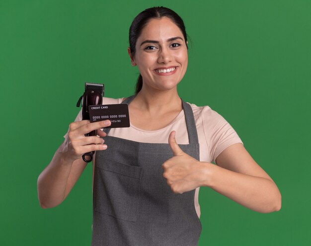 Jeune belle femme coiffeur en tablier tenant tondeuse et carte de crédit à l'avant avec le sourire sur le visage montrant les pouces vers le haut debout sur le mur vert