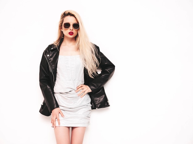 Jeune belle femme confiante en robe argentée de soirée d'été à la mode Sexy femme blonde insouciante posant près d'un mur blanc en studio Modèle en veste de motard en cuir noir et lèvres rouges