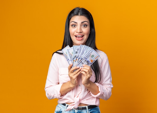 Jeune belle femme dans des vêtements décontractés tenant de l'argent, heureux et positif souriant joyeusement debout sur le mur orange