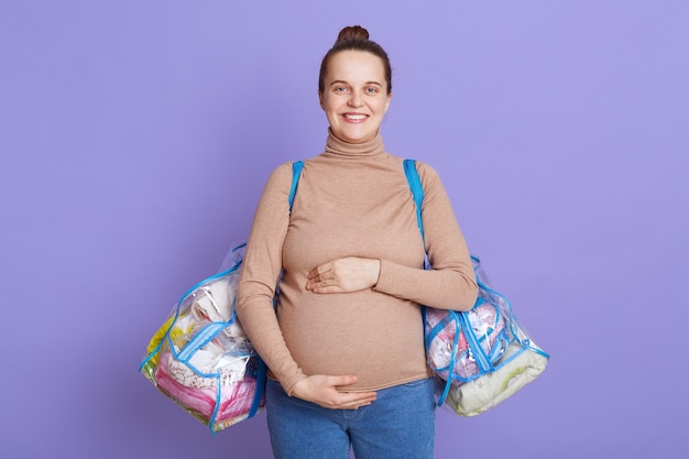 Jeune belle femme enceinte debout isolée sur mur lilas, toucher son ventre, tenant des sacs avec des trucs pour la maison de maternité.