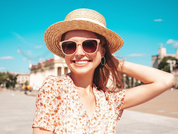 Jeune belle femme hipster souriante dans des vêtements d'été à la mode Sexy femme insouciante posant sur le fond de la rue au coucher du soleil Modèle positif à l'extérieur Gai et heureux en lunettes de soleil et chapeau