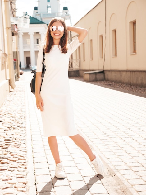 Jeune belle femme hipster souriante dans des vêtements d'été à la mode Sexy femme insouciante posant sur le fond de la rue au coucher du soleil Modèle positif à l'extérieur Gai et heureux en lunettes de soleil