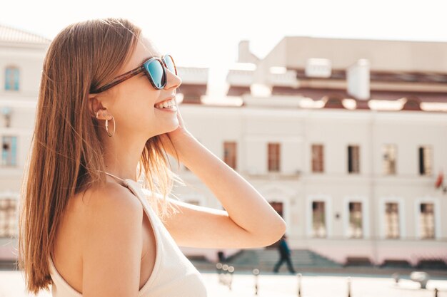 Jeune belle femme hipster souriante dans des vêtements d'été à la mode Sexy femme insouciante posant sur le fond de la rue au coucher du soleil Modèle positif à l'extérieur Gai et heureux en lunettes de soleil