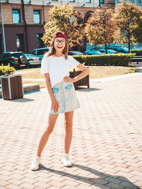 Jeune belle femme hipster souriante dans des vêtements d'été à la mode Sexy femme insouciante posant sur le fond de la rue en casquette au coucher du soleil Modèle positif à l'extérieur Gai et heureux en lunettes de soleil