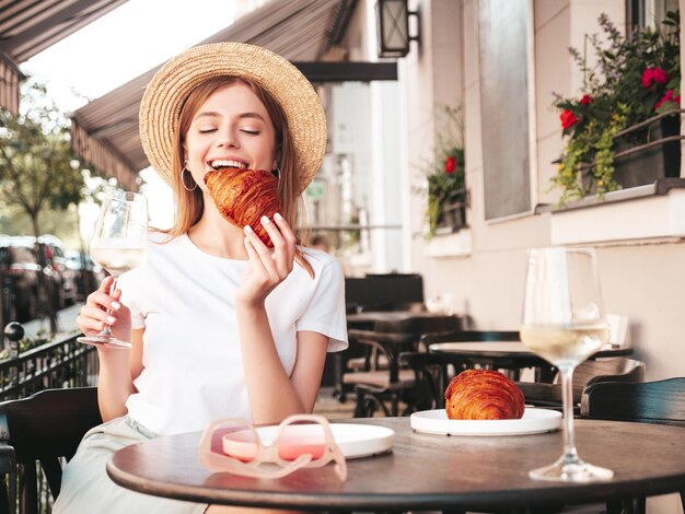 Jeune belle femme hipster souriante dans des vêtements d'été à la modeFemme insouciante posant au café-véranda dans la rueModèle positif buvant du vin blancProfitant des vacances