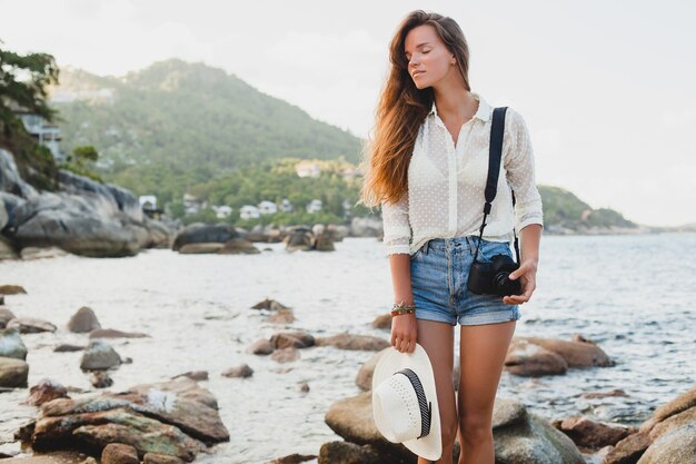 Jeune belle femme hipster en vacances d'été en Asie, détente sur la plage tropicale, appareil photo numérique, style boho décontracté, paysage de mer, corps bronzé mince, voyage seul