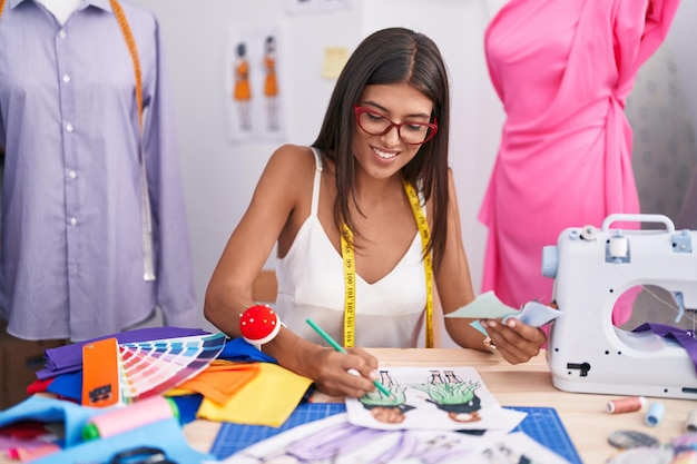 Jeune belle femme hispanique tailleur souriant confiant dessinant la conception de vêtements à la boutique de tailleur