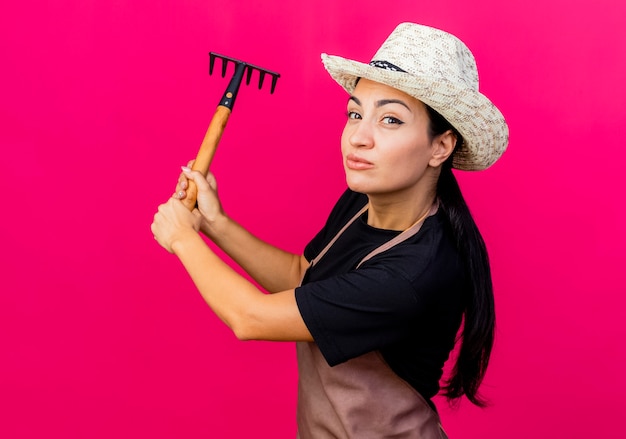 Jeune belle femme jardinier en tablier et chapeau balançant mini râteau souriant debout sur un mur rose