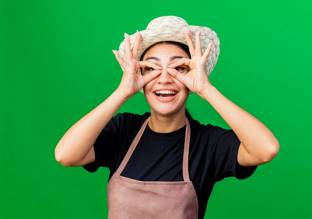 Photo gratuite jeune belle femme jardinier en tablier et chapeau faisant un geste binoculaire avec les doigts regardant à travers les doigts debout sur fond vert