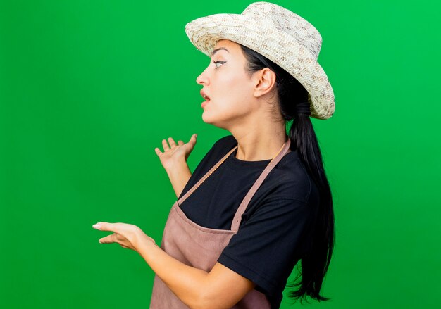 Jeune belle femme jardinier en tablier et chapeau présentant quelque chose avec les mains derrière elle