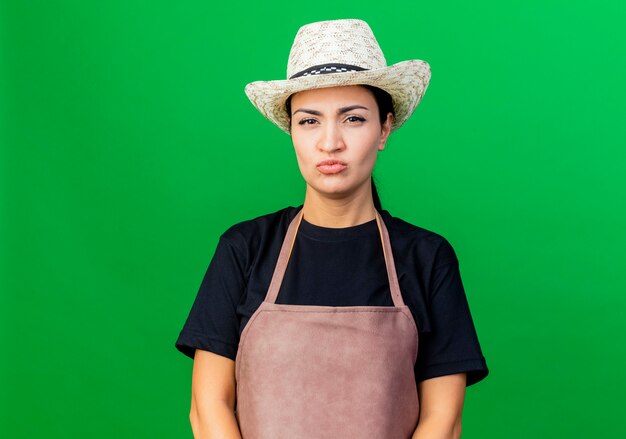 Jeune belle femme jardinier en tablier et chapeau regardant à l'avant avec un visage sérieux d'être mécontent debout sur mur vert