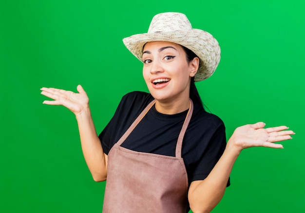 Jeune belle femme jardinier en tablier et chapeau souriant confus écartant les bras sur les côtés