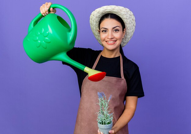 Jeune belle femme jardinier en tablier et chapeau tenant arrosoir et plante en pot à l'avant souriant debout sur le mur bleu
