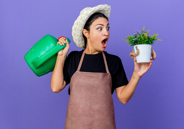 Jeune belle femme jardinier en tablier et chapeau tenant arrosoir et plante en pot étant étonné et surpris debout sur le mur bleu