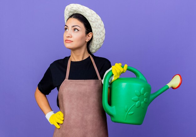 Jeune belle femme jardinier en tablier et chapeau tenant arrosoir à la recherche de côté avec une expression pensive sur le visage debout sur un mur bleu