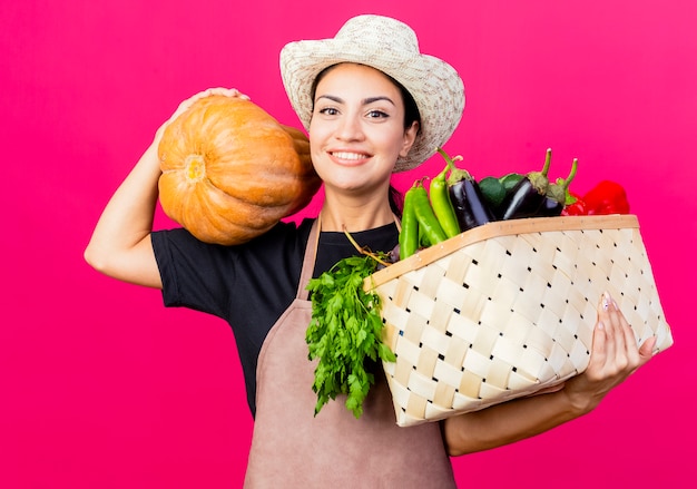 Jeune belle femme jardinier en tablier et chapeau tenant une caisse pleine de légumes et de citrouille lookign à la caméra avec un visage heureux