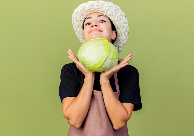 Jeune belle femme jardinier en tablier et chapeau tenant le chou à l'avant souriant avec un visage heureux debout sur un mur vert clair