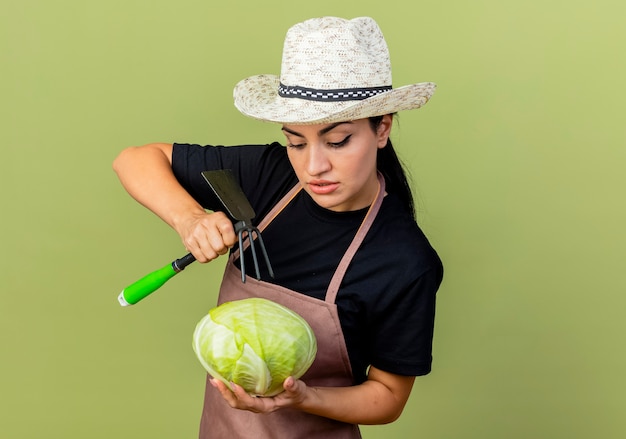 Jeune belle femme jardinier en tablier et chapeau tenant le chou et la pioche le regardant avec un visage sérieux debout sur un mur vert clair