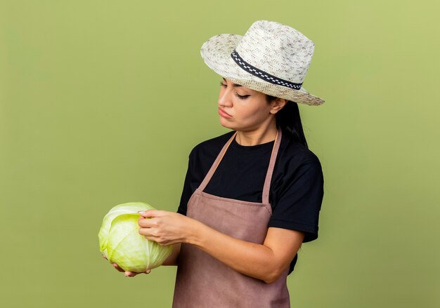 Jeune belle femme jardinier en tablier et chapeau tenant chou en regardant avec un visage sérieux debout sur un mur vert clair
