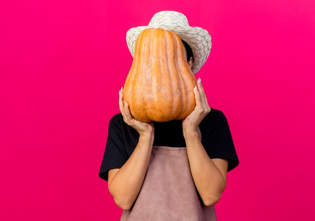 Jeune belle femme jardinier en tablier et chapeau tenant la citrouille cachant son visage debout sur un mur rose