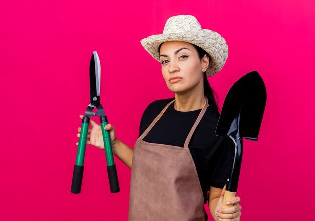 Jeune belle femme jardinier en tablier et chapeau tenant un coupe-haie et une pelle à l'avant avec un visage sérieux debout sur un mur rose