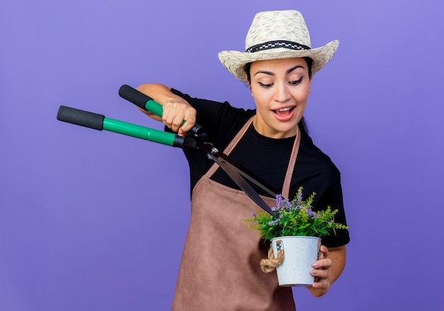 Jeune belle femme jardinier en tablier et chapeau tenant un coupe-haie et plante en pot souriant debout sur un mur bleu