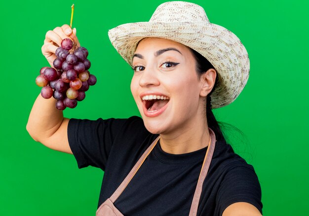 Jeune belle femme jardinier en tablier et chapeau tenant grappe de raisin souriant joyeusement