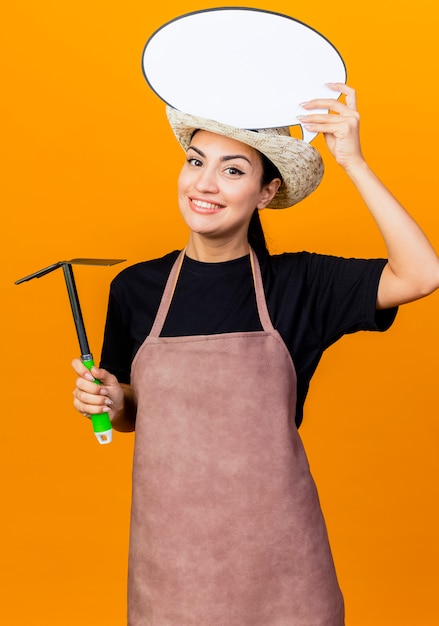 Jeune Belle Femme Jardinier En Tablier Et Chapeau Tenant Mattock Et Signe De Bulle De Discours Vide Sur La Tête Souriant Debout Sur Le Mur Orange