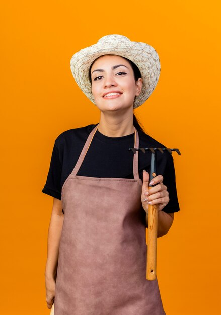 Jeune belle femme jardinier en tablier et chapeau tenant un mini râteau à l'avant souriant avec un visage heureux debout sur un mur orange