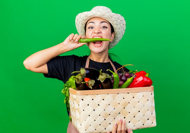 Jeune belle femme jardinier en tablier et chapeau tenant un panier plein de légumes mordant le piment vert debout sur le mur vert