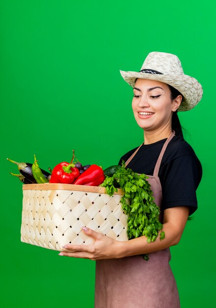 Jeune belle femme jardinier en tablier et chapeau tenant un panier plein de légumes souriant avec un visage heureux debout sur un mur vert