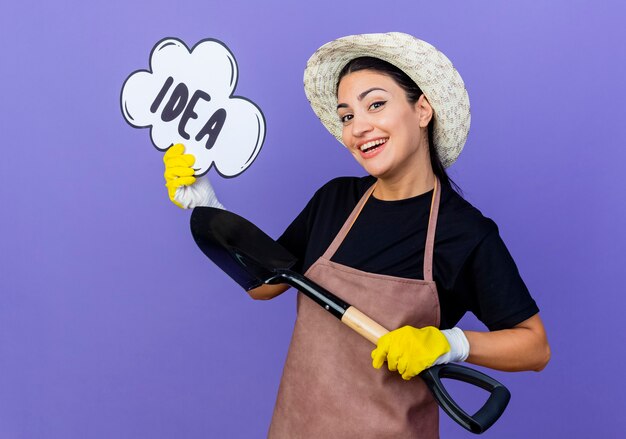 Jeune belle femme jardinier en tablier et chapeau tenant la pelle et le signe de la bulle de la parole avec l'idée de mot souriant confiant debout sur le mur bleu