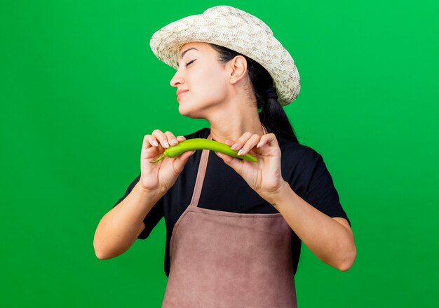 Jeune belle femme jardinier en tablier et chapeau tenant le piment vert avec les yeux fermés