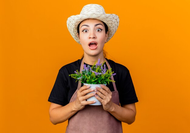 Jeune belle femme jardinier en tablier et chapeau tenant une plante en pot à l'avant d'être surpris debout sur un mur orange