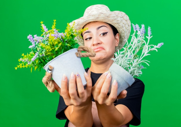Photo gratuite jeune belle femme jardinier en tablier et chapeau tenant des plantes en pot à la confusion et mécontent debout sur mur vert