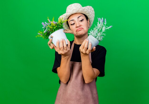 Jeune belle femme jardinier en tablier et chapeau tenant des plantes en pot à la confusion et mécontent