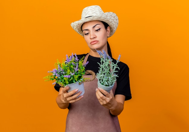 Jeune belle femme jardinier en tablier et chapeau tenant des plantes en pot les regardant confus debout sur un mur orange