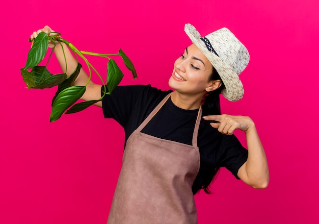 Jeune Belle Femme Jardinier En Tablier Et Chapeau Tenant Pointign Plante Avec Index à Elle Souriant Debout Sur Le Mur Rose