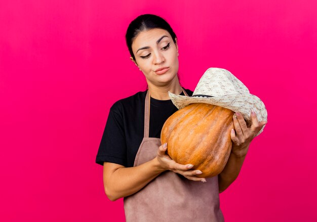 Jeune belle femme jardinier en tablier holding pumpking avec hat lookign avec une expression sceptique