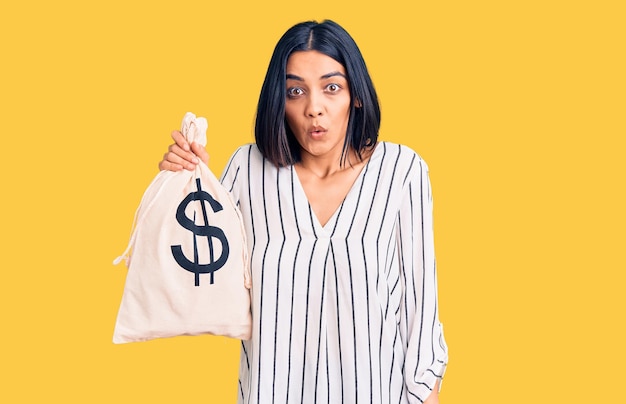 Photo gratuite jeune belle femme latine tenant un sac d'argent avec le symbole du dollar effrayé et étonné avec la bouche ouverte pour le visage d'incrédulité surprise