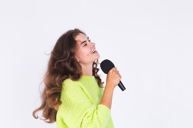 Jeune belle femme avec un maquillage léger de taches de rousseur en pull sur un mur blanc avec un microphone chantant heureux en mouvement