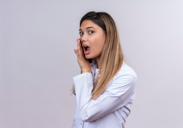 Jeune belle femme médecin vêtu d'un blouse blanche avec stéthoscope disant un secret avec les mains près de la bouche