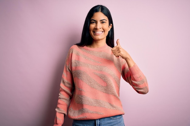 Jeune belle femme de mode hispanique portant un chandail imprimé animal sur fond rose faisant un geste de pouce levé heureux avec la main approuvant l'expression regardant la caméra montrant le succès