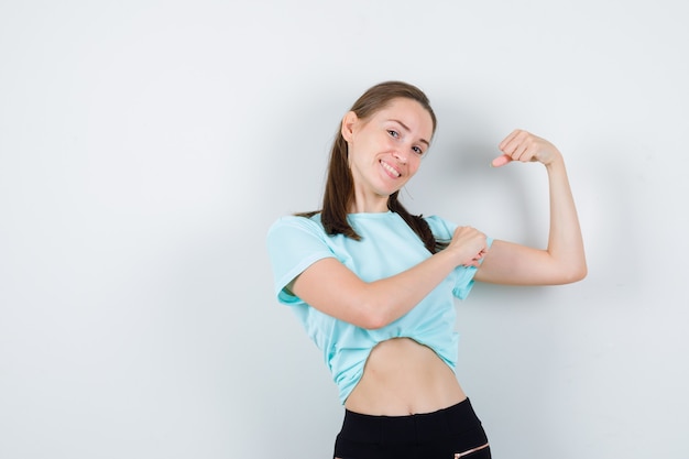 Jeune belle femme montrant les muscles du bras, avec la main sur le bras en t-shirt et l'air fier, vue de face.