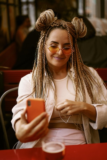 Jeune belle femme multiraciale avec une coiffure afro blonde tresses zizi sur le café de la rue avec un casque. lunettes de soleil jaunes, maquillage lumineux, style hippie. appel vidéo, travail à l'extérieur de la maison