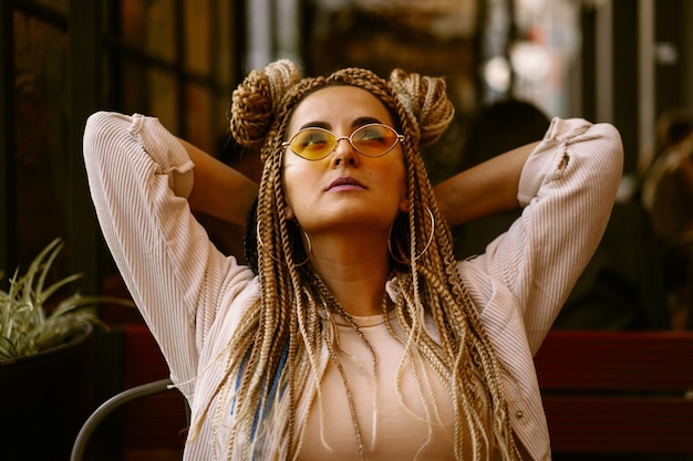Jeune belle femme multiraciale avec une coiffure afro blonde tresses zizi sur le café de la rue. lunettes de soleil jaunes, maquillage lumineux, style hippie